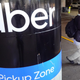 A Pole with the Uber logo and text under it saying "pickup zone." A man with a bag walks by holding his phone, waiting for his Uber pickup.